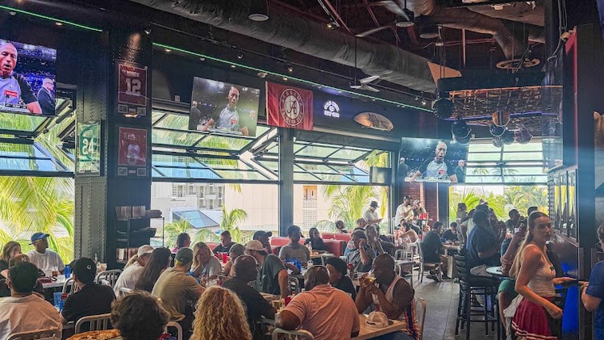 Packed crowd watching sports at Skybox Taphouse Waikiki with palm trees and open-air seating